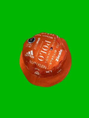 Orange bucket hat med logotyper - Cool orange bucket hat med tryckta logotyper från flera kända märken som Adidas, Polo, Calvin Klein och Guess. Hatten har en klassisk form med bred skärm och är tillverkad i ett lätt tygmaterial. Perfekt för dig som vill sticka ut och visa din stil.
