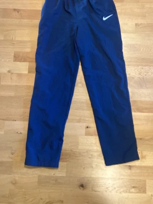 Blå träningsbyxor från Nike - Snygga blå träningsbyxor från Nike med klassisk vit logga på vänster lår. Byxorna har rak passform och resår i midjan för skön komfort. Perfekta för gymmet eller löprundan. Tillverkade i lätt och funktionellt material.