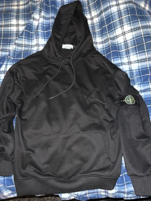 Svart hoodie från Stone Island - Snygg svart hoodie från Stone Island med klassisk patch på vänster ärm. Hoodien har luva med dragsko och en stor magficka framtill. Tillverkad i mjukt bomullsmaterial som är skönt att bära. Perfekt för dig som gillar streetwear och vill ha en stilren look.