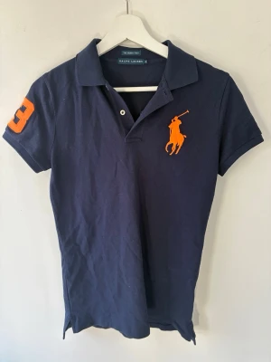 Marinblå pikétröja från Ralph Lauren - Klassisk marinblå pikétröja från Ralph Lauren med orange logga på bröstet och en stor orange trea på ärmen. Tröjan har krage, korta ärmar och knäppning framtill. Det står att storleken är M men skulle säga att den sitter mer åt S hållet.