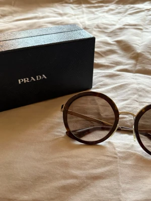 Runda solglasögon från Prada - Snygga runda solglasögon från Prada med brunmelerad båge och tunna metallskalmar. Klassisk och trendig design som ger en cool retrovibe. Kommer med originaletui från Prada.