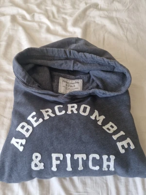 Abercrombie & Fitch Hoodie - Säljer en mörkgrå hoodie från Abercrombie & Fitch med vit logotyp på bröstet. Hoodien har huva, långa ärmar och en stor magficka. Materialet är mjukt och bekvämt, perfekt för chill dagar. Klassisk och stilren design som passar till jeans eller leggings.