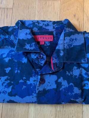 Blå camouflage skjorta från Vistula - Säljer en blå camouflage-mönstrad skjorta från Vistula. Skjortan har klassisk krage, knappar framtill och coola röda sömmar vid kragen. Perfekt för dig som vill sticka ut med en unik look.