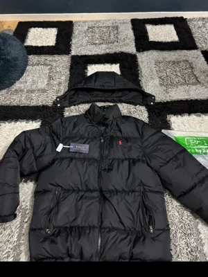 Svart pufferjacka från Polo Ralph Lauren - Snygg svart pufferjacka från Polo Ralph Lauren med avtagbar huva och dragkedja framtill. Jackan har två framfickor med dragkedja och den klassiska röda Polo-loggan broderad på bröstet. Perfekt för kalla vinterdagar och riktigt stilren look.