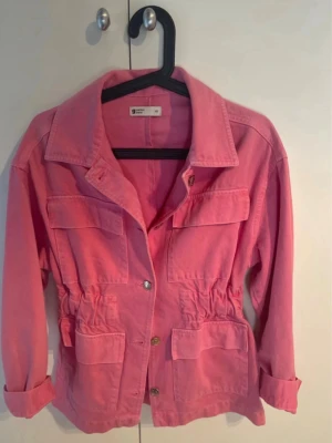 Rosa jeansjacka från Gina Tricot - Snygg rosa jeansjacka från Gina Tricot med fyra stora fickor framtill, knappar och markerad midja med resår. Jackan har klassisk krage och långa ärmar som kan vikas upp för en avslappnad look. Perfekt för dig som vill sticka ut med färg.