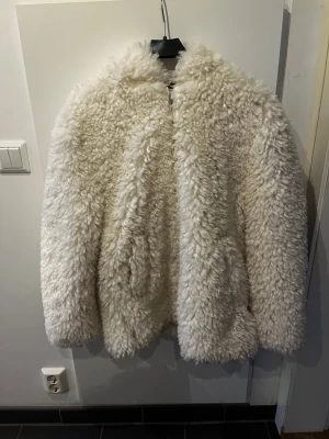 Vit fluffig teddyjacka med huva - Säljer en supermjuk och fluffig teddyjacka i vit färg. Jackan har en oversized passform och en stor huva som ger extra mysfaktor, har en kort slit på båda sidor. Perfekt för dig som vill ha en trendig och varm jacka till hösten och vintern. Materialet är fuskpäls som känns riktigt gosigt. Jättefin och cool men var inte som jag tänkte så endast testad få korta stunder. 