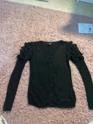 Svart kofta med volangärmar H&M - Snygg svart kofta från H&M med knappar framtill och unika volangdetaljer på axlarna. Långärmad och i mjukt, tunt material som passar perfekt till lager på lager. V-ringad och enkel att styla till olika outfits.