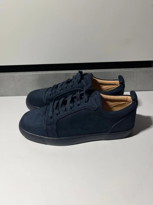 Mörkblå sneakers från Christian Louboutin - Stilrena mörkblå sneakers i mocka från Christian Louboutin med klassisk röd sula och diskret logga undertill. Lågt skaft, matchande mörkblå snörning och beige innerfoder. Perfekta för dig som gillar exklusiv och clean stil. Helt nya