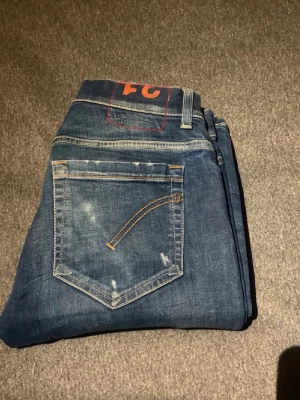 Mörkblå  jeans med slitningar - Säljer ett par mörkblå skinny jeans med slitningar och distressed detaljer på benen. Jeansen har klassisk femficksmodell, orange sömmar och en cool FC-logga i midjan. Perfekta för en avslappnad och trendig look.