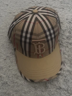 Beige Burberry keps med rutmönster - Säljer en snygg beige keps från Burberry med det klassiska rutmönstret i svart, vitt och rött. Framtill finns ett broderat Burberry-monogram i beige. Justerbar rem bak och böjd skärm. Perfekt för dig som vill ha en ikonisk accessoar med lyxig känsla.