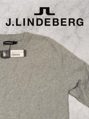 Grå stickad tröja från J.Lindeberg - Snygg grå stickad tröja från J.Lindeberg i bomull med strukturerat mönster. Tröjan har rund halsringning och ribbade muddar vid ärmslut och nederkant. Perfekt för en stilren och avslappnad look.