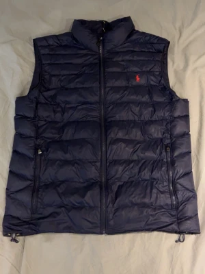 Mörkblå dunväst från Ralph Lauren - Snygg mörkblå dunväst från Ralph Lauren med hög krage och dragkedja framtill. Västen har två fickor med dragkedja och den klassiska röda Polo-loggan på bröstet. Perfekt för lager-på-lager och håller dig varm under kyliga dagar. Storlek XXL passar som L-XL, skickas samma dag eller dagen efter köp