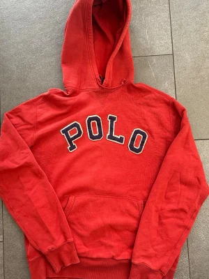 Röd hoodie från Ralph Lauren - Herr M - Snygg röd hoodie från Polo Ralph Lauren med stor POLO-text i vitt och blått på bröstet. Tröjan är i okej skick men har några skavanker vid muddarna (se bilder) samt en mindre fläck på fickan. Nypris ligger runt 2500kr+