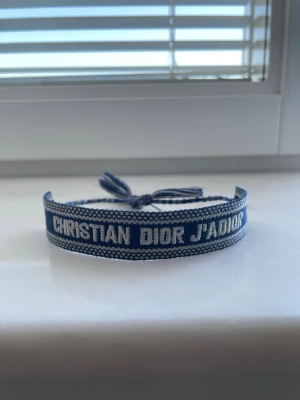 Blått armband ifrån Dior - Snyggt blått tygarmband från Christian Dior med texten 'CHRISTIAN DIOR J'ADIOR' broderad i vitt. Armbandet har justerbar knytning med tofsar i ändarna och en cool, avslappnad look. Perfekt accessoar för att lyfta din outfit.