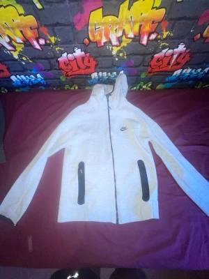 Ljusgrå hoodie från Nike - Säljer en ljusgrå hoodie från Nike med dragkedja framtill och två svarta dragkedjeförsedda fickor. Hoodien har Nike-logga på bröstet och är tillverkad i mjukt sweatshirtmaterial. Perfekt för en sportig och avslappnad stil.