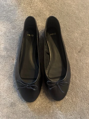 Svarta ballerinaskor från Mango - Svarta ballerinaskor från Mango med rund tå och liten rosett framtill. Skorna är i ett slätt material. Endast använda två gånger. Säljer då de är lite för stora för mig.💋original pris: 599kr