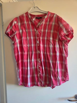 Rutig rosa blus från Lindex, stl 48 - Säljer en luftig rosa blus från Lindex med vitt och blått rutmönster. Blusen har korta ärmar, knappar framtill och en avslappnad passform. Perfekt till jeans eller shorts för en casual look.