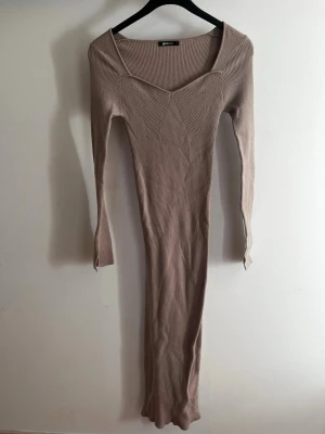 Beige ribbad klänning Gina Tricot - Lång beige ribbad klänning från Gina Tricot med lång ärm och v-ringad halsringning. Klänningen har en figurnära passform och är tillverkad i ett stretchigt material som sitter snyggt på kroppen. Perfekt för dig som gillar stilrena och enkla plagg. Säljes för jag tyvärr tycker den är för kort för mig som är 173, den passar nog på andra som också är längre men jag vill att den ska gå ner över hela benen och det gör den inte.