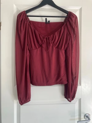 Vinröd omlottblus  - Säljer en vinröd blus från COS med omlottdetalj och knyt framtill. Blusen har långärmade puffiga ärmar och en mjuk, slät yta. Modellen är croppad och har en snygg v-ringning med rynkade detaljer vid bysten. Perfekt för dig som gillar stilrena och trendiga plagg.