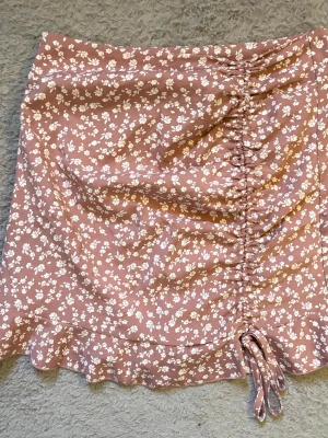 Rosa blommig kortkjol med snörning - Supersöt kortkjol i ljusrosa med vitt blommönster och snörning framtill. Kjolen har volangkant nedtill och är gjord i ett mjukt, lätt material som känns skönt mot huden. Perfekt för dig som gillar romantisk och trendig stil.