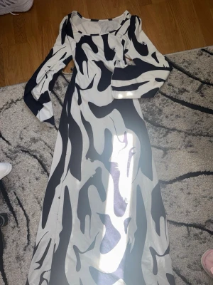 Lång svartvit mönstrad klänning - Snygg långklänning med svartvitt abstrakt mönster och långa ärmar. Klänningen har snörning i midjan för en cool detalj och markerad siluett. Perfekt för dig som vill sticka ut med en unik look.