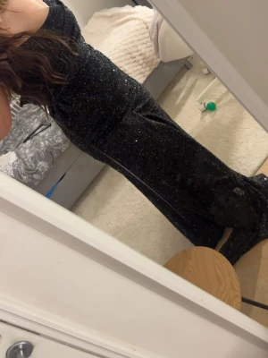 Svart glittrig långklänning - Säljer en svart långklänning med glittrigt tyg och smala axelband. Klänningen har en figurnära passform och är perfekt för speciella tillfällen. Materialet känns mjukt och följsamt mot kroppen och ger en elegant look. Den har även typ öppen rugg där man kan spänna åt, minns tyvärr inte vart jag köpt den