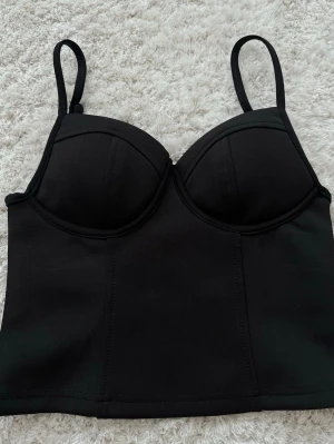 Svart korset topp - Snygg svart korset topp med vadderade kupor och justerbara axelband. Lång modell som ger extra stöd och formar midjan. Tillverkad i stretchigt material för en smickrande siluett. Perfekt under tighta toppar eller klänningar.