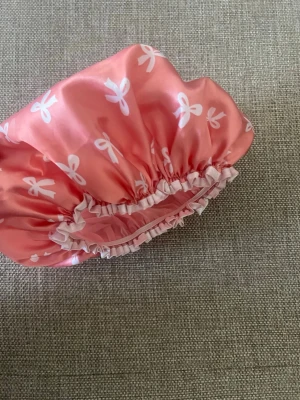 Rosa satin duschmössa med rosettprint - Söt duschmössa i glansig rosa satin med vita rosetter som mönster. Mössan har elastisk kant med volangdetalj för att sitta bekvämt på huvudet. Perfekt för att skydda håret vid dusch eller bad.💗💕💞
