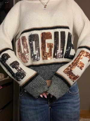 Vit stickad tröja med paljetter - Trendig vit stickad tröja med stora paljettbokstäver i svart, koppar och silver som stavar 'VOGUE'. Tröjan har breda muddar i grått och svart samt avslappnad passform och rund hals. Perfekt för dig som vill sticka ut med en unik och cool look.