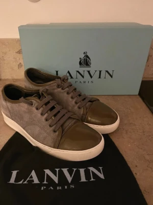 Gråa Lanvin sneakers med gröna detaljer - Snygga sneakers från Lanvin i grå mocka med gröna lackade tåpartier och matchande gröna skosnören. Skorna har vit sula och klassisk låg profil. Perfekta för dig som vill ha en stilren men unik sneaker från ett lyxigt märke.