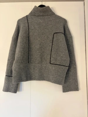 Grå stickad polotröja med detaljer - Säljer en grå stickad polotröja med oversized passform och svarta kontrastsömmar. Tröjan har ribbade muddar, hög krage och en unik asymmetrisk design med markerade sömmar. Perfekt för dig som gillar en modern och avslappnad stil.