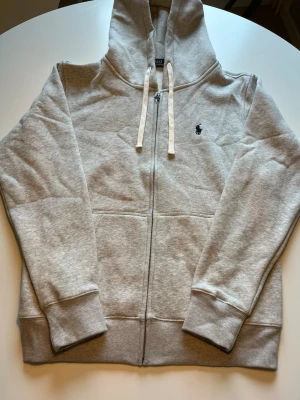 Polo Ralph Lauren zip hoodie - Snygg grå hoodie från Polo Ralph Lauren med klassisk broderad logga på bröstet. Tröjan har huva med vita snören, hel dragkedja och ribbade muddar. Perfekt för chill dagar och enkel att matcha med jeans eller joggers. Perfekt skick!