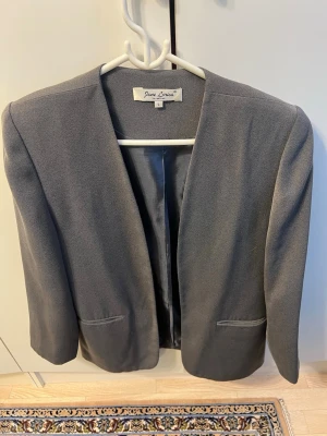 Grå kavaj från Jane Lorisa storlek L - Stilren grå kavaj från Jane Lorisa med öppen front utan knappar och två fickor framtill. Kavajen har en rak passform och är helfodrad för extra komfort. Perfekt för en clean och modern look.