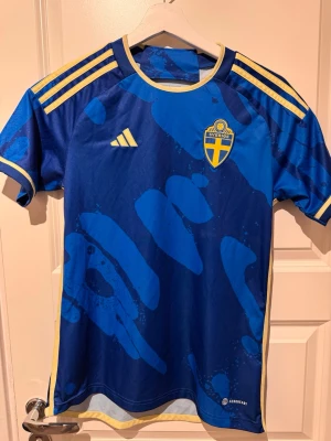 Blå Sverige fotbollströja Adidas - Säljer en äkta blå fotbollströja från Adidas med Sveriges landslagsemblem på bröstet. Tröjan har gula detaljer, korta ärmar och ett unikt blått mönster. Tillverkad i lätt funktionsmaterial med Aeroready-teknologi för bästa komfort.