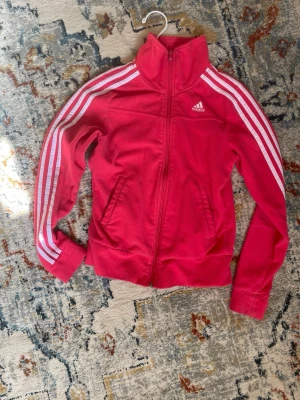 Röd Adidas  - Säljer en klassisk röd Adidas träningsjacka med vita ränder längs ärmarna och dragkedja framtill. Jackan har hög krage, två fickor och Adidas-logga på bröstet. Perfekt till träning eller sportig vardagsstil.