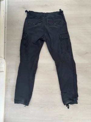 Svarta cargobyxor från H&M L.O.G.G. - Säljer ett par svarta cargobyxor från H&M L.O.G.G. med flera praktiska fickor på sidorna och baktill. Byxorna har dragsko vid bensluten och är tillverkade i ett slitstarkt bomullsmaterial. Perfekta för en avslappnad och cool stil.