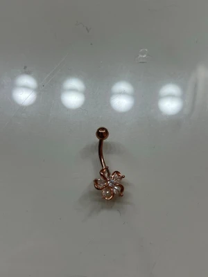 Navelpiercing med blomma i roséguld - Snygg navelpiercing i roséguld med en blomformad detalj dekorerad med små glittrande stenar. Blomman har fem kronblad och en gnistrande mitt, vilket ger ett elegant och trendigt intryck. Perfekt accessoar för att lyfta din stil.
