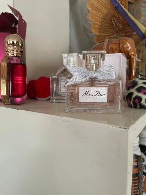 Miss Dior Parfum med silverrosett - Miss Dior Parfum från Dior i en elegant, rektangulär glasflaska med ljusrosa doftvätska. Flaskan har en dekorativ silverfärgad rosett vid halsen och en stilren etikett framtill. Perfekt för dig som gillar lyxiga och klassiska parfymer.