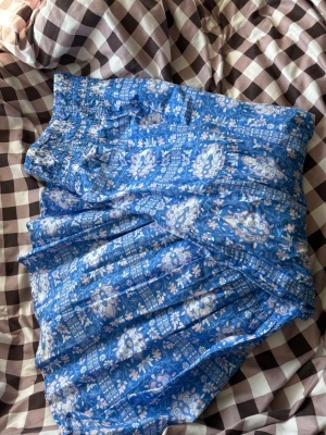 Fint somrigt sett med kjol och topp - Säljer en blå långkjol med vitt och ljusblått blommönster. Kjolen har bred resår i midjan för skön passform och är gjord i ett lätt och luftigt material, perfekt för varma dagar. Passar dig som vill ha något färgglatt i garderoben. Det är ett sett med en topp som är som kjolen, man kan ta av banden på toppen för att använda den utan band eller som en kjol t.ex. helt nytt. Strl 170 men passar S och M