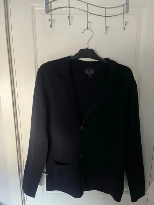 Mörkblå stickad kofta från H&M - Stilren mörkblå kofta/ stickad kavaj från H&M med klassisk krage och två framfickor. Kavajen har tre knappar framtill och är tillverkad i ett mjukt, stickat material som ger en avslappnad men ändå dressad look. Perfekt för dig som vill ha en enkel och snygg kavaj.