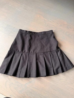 Svart plisserad kortkjol - Snygg svart kortkjol med breda plisserade veck. Kjolen har en klassisk A-linjeform och är tillverkad i ett mjukt bomullsliknande material. Perfekt att matcha med en crop top eller hoodie för en trendig look.