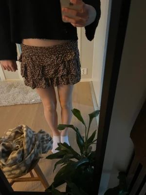 Brun blommig volangkjol - Supersöt brun kortkjol med små vita blommor och volangdetaljer. Kjolen har en flowy passform och är perfekt för vår och sommar. Tillverkad i ett lätt och mjukt material som känns skönt mot huden.