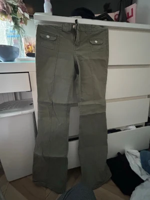 Olivgröna bootcut jeans med fickor - Snygga olivgröna jeans med bootcut-modell och två framfickor med lock och knapp. Perfekta för dig som gillar en avslappnad men trendig stil. Jeansen är i storlek 38 och väldigt fint skick