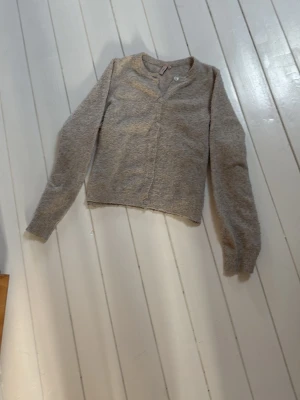 Beige kofta från Nelly med rund hals - Mysig beige kofta från Nelly med rund halsringning och knäppning framtill. Tillverkad i ett mjukt, luddigt material som känns skönt mot huden. Perfekt att slänga över en t-shirt eller linne för en avslappnad look.