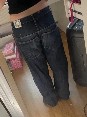 Baggy mörkblå jeans - Säljer ett par baggy mörkblå jeans med raka ben och stora bakfickor. Jeansen har en vit patch med en dödskalle på linningen bak och klassisk femficksdesign. Väldigt skönt material för att vara jeans. Finns lite slitning på benen längst ner
