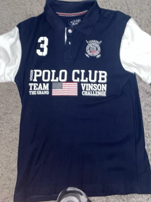 Mörkblå piké från Vinson Polo Club - Mörkblå pikétröja från Vinson Polo Club med vita kontrastärmar och tryck framtill. Klassisk krage, knappar vid halsen och detaljer som amerikansk flagga och broderad logga. Perfekt för dig som gillar sportig stil.