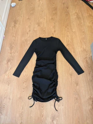 Svart ribbad klänning med snörning - Trendig svart ribbad klänning med långa ärmar och snörningsdetaljer på båda sidorna av kjolen. Klänningen har en kroppsnära passform och är tillverkad i ett mjukt stretchigt material. Perfekt för dig som gillar en stilren look med coola detaljer.