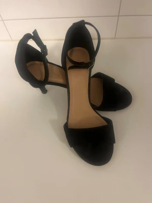 Svarta pumps med ankelrem - Stilrena svarta pumps med öppen tå och smal klack. Skorna har en bred rem över tårna och justerbar ankelrem med spänne. Insidan är beige och materialet ser ut att vara mockaimitation. Perfekta för dig som gillar klassisk och enkel stil.