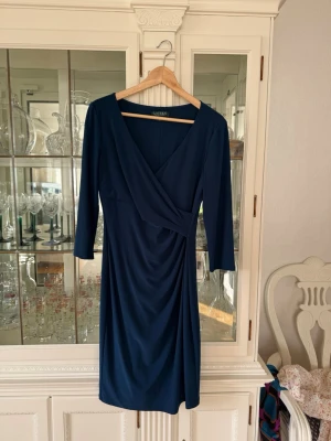 Marinblå omlottklänning Lauren Ralph Lauren - Snygg marinblå omlottklänning från Lauren Ralph Lauren med v-ringning och trekvartsärm. Klänningen har draperad midja och elegant, enkel design som ger en smickrande siluett. Perfekt för dig som gillar klassisk stil.