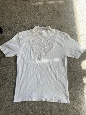 Vit topp med spetsdetalj i halsen - Säljer en vit topp med korta ärmar och en snygg spetsdetalj framtill vid halsen. Spetsen har ett elegant mönster och ger toppen en unik look. Perfekt för dig som vill ha något extra till din outfit.
